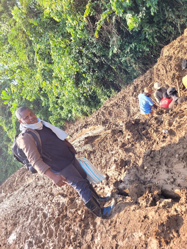 Photograph showing a muddy outdoor area with three people engaged in digging or excavation activities near dense green foliage. One person stands in the foreground wearing boots and holding a bag, while two others work closely together in the mud.