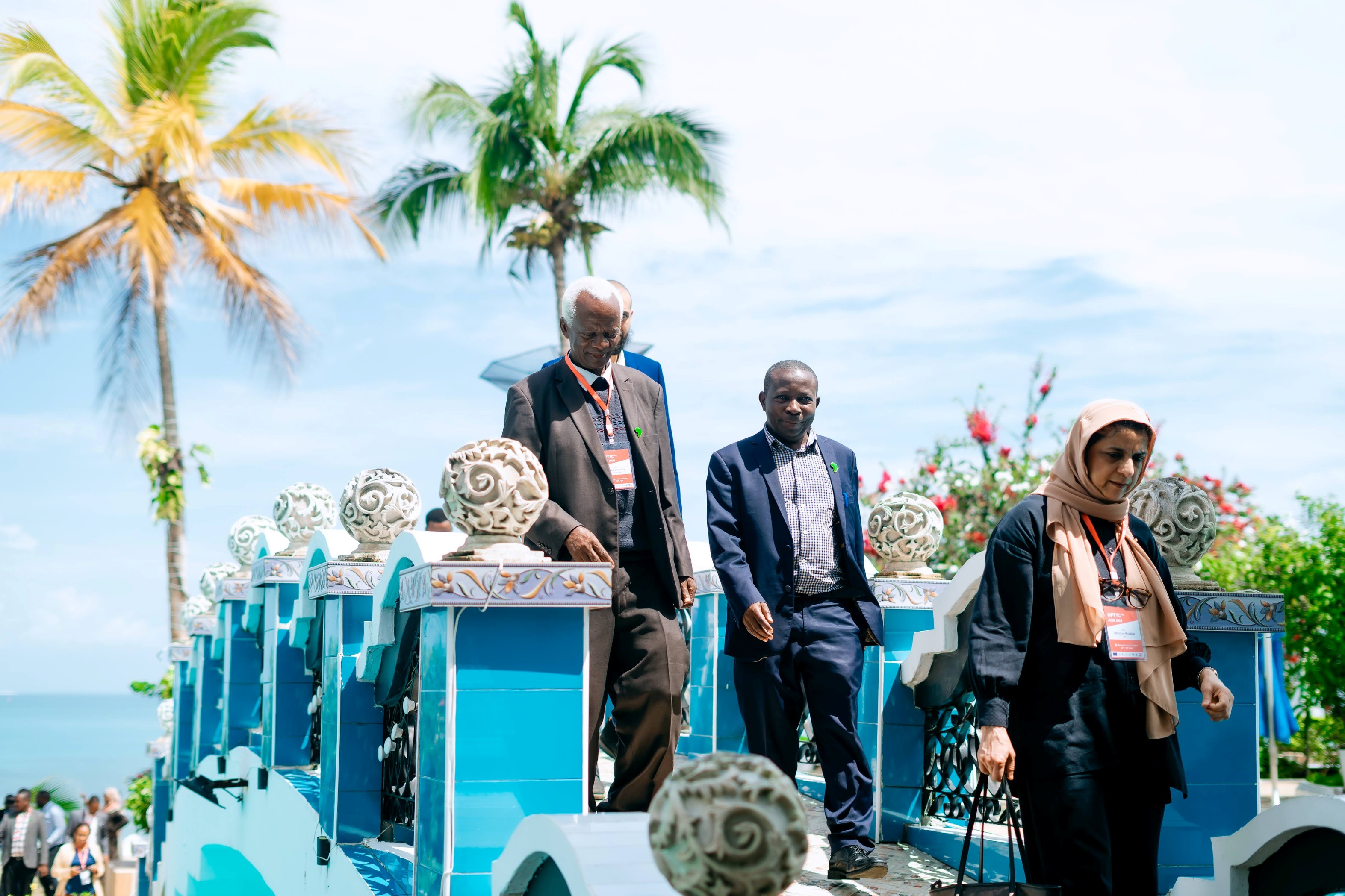 Photograph showing three people walking down a blue decorative bridge with ornate white spherical finials, set against a backdrop of palm trees and a partly cloudy sky. The scene suggests a formal or professional event, indicated by attendees wearing badges and business attire.